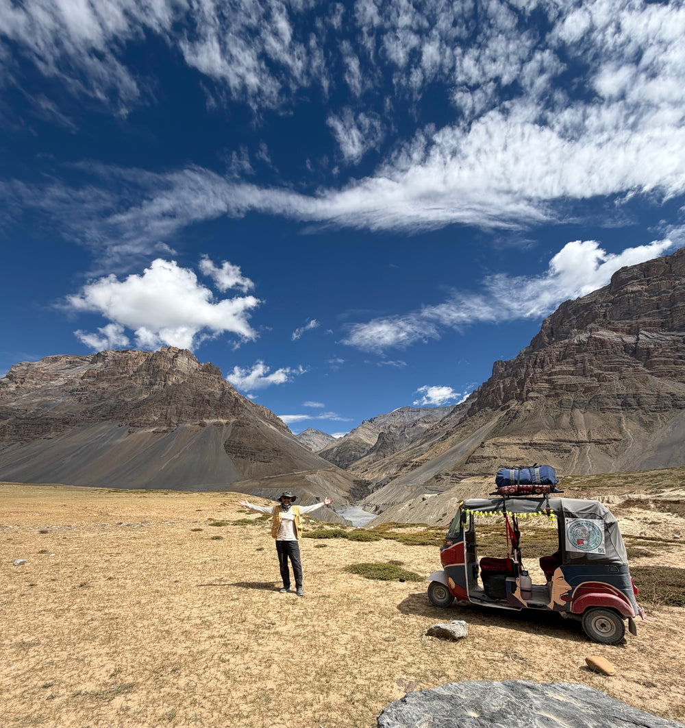 A person stands in a barren mountain valley with arms outstretched under a bright blue sky with white clouds, beside a small red and gray three-wheeler vehicle loaded with gear.