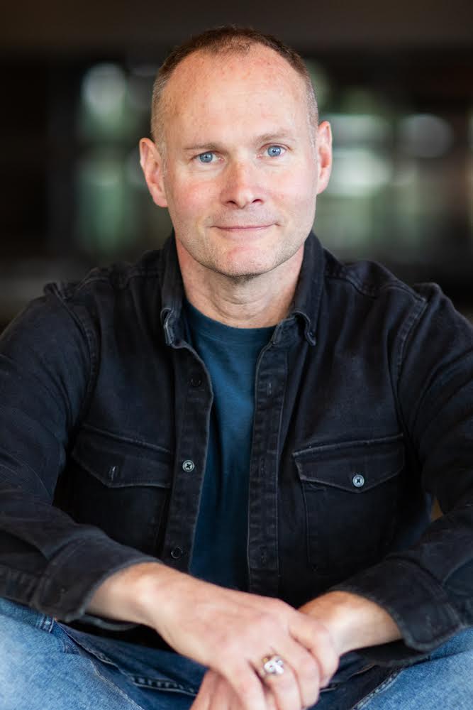A man wearing a black denim button-up shirt over a dark teal t-shirt and blue jeans, sitting with his hands crossed. He is wearing a silver ring on his left hand.