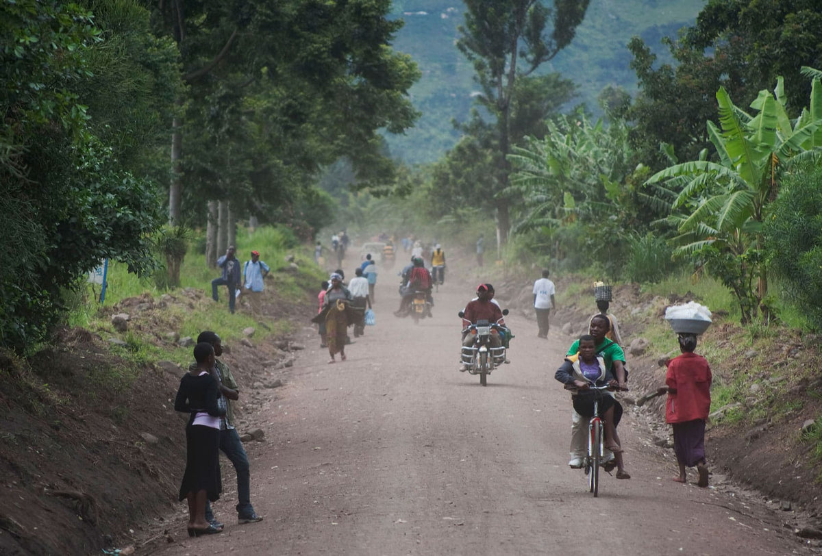 A dusty dirt road through a lush green rural landscape, with several people riding bicycles and motorcycles and a few walking. Trees, grass, and banana plants line both sides of the road.