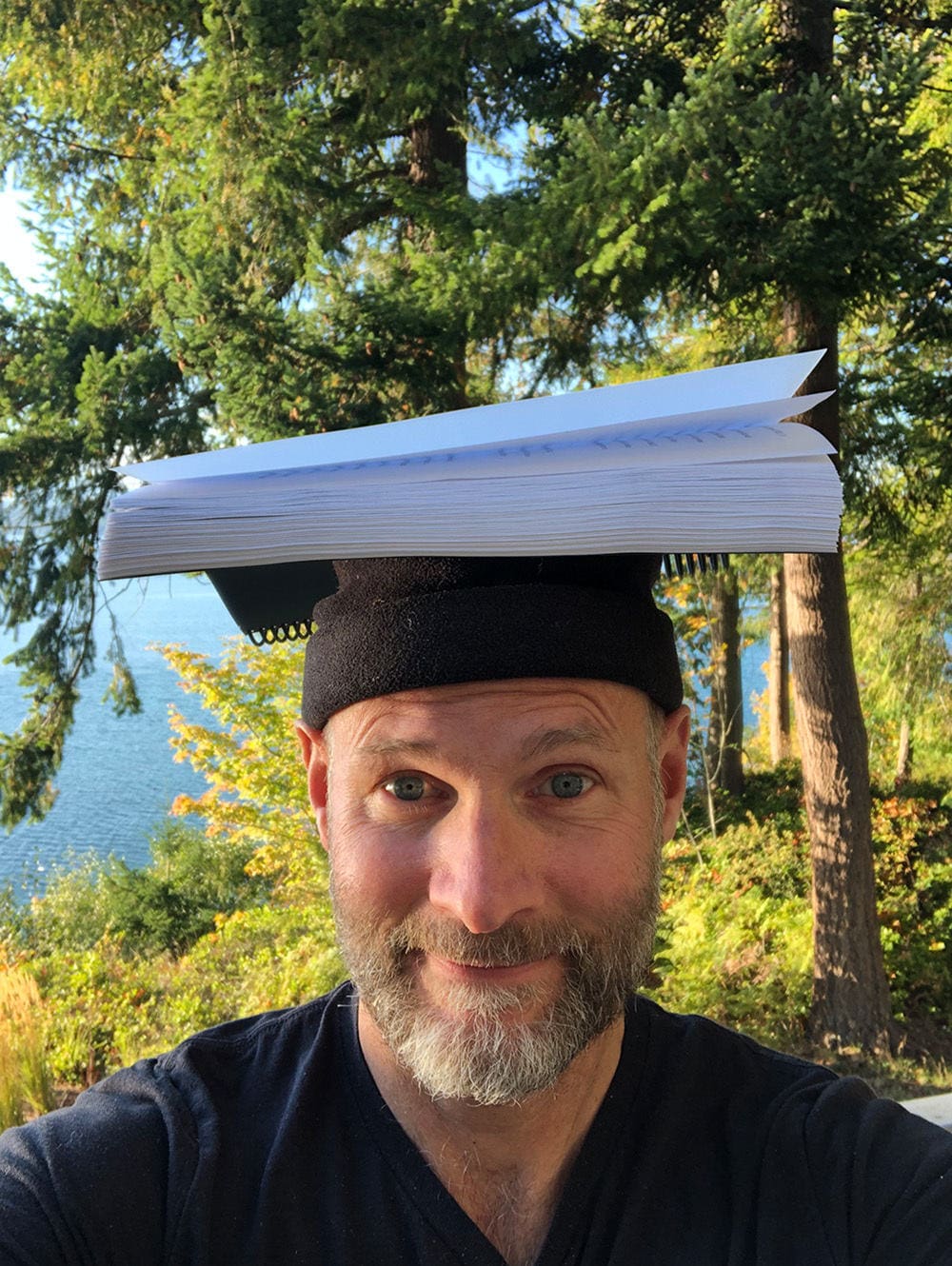 A man outdoors with trees and water in the background, wearing a black cap with a large white notebook balanced on top like a hat.