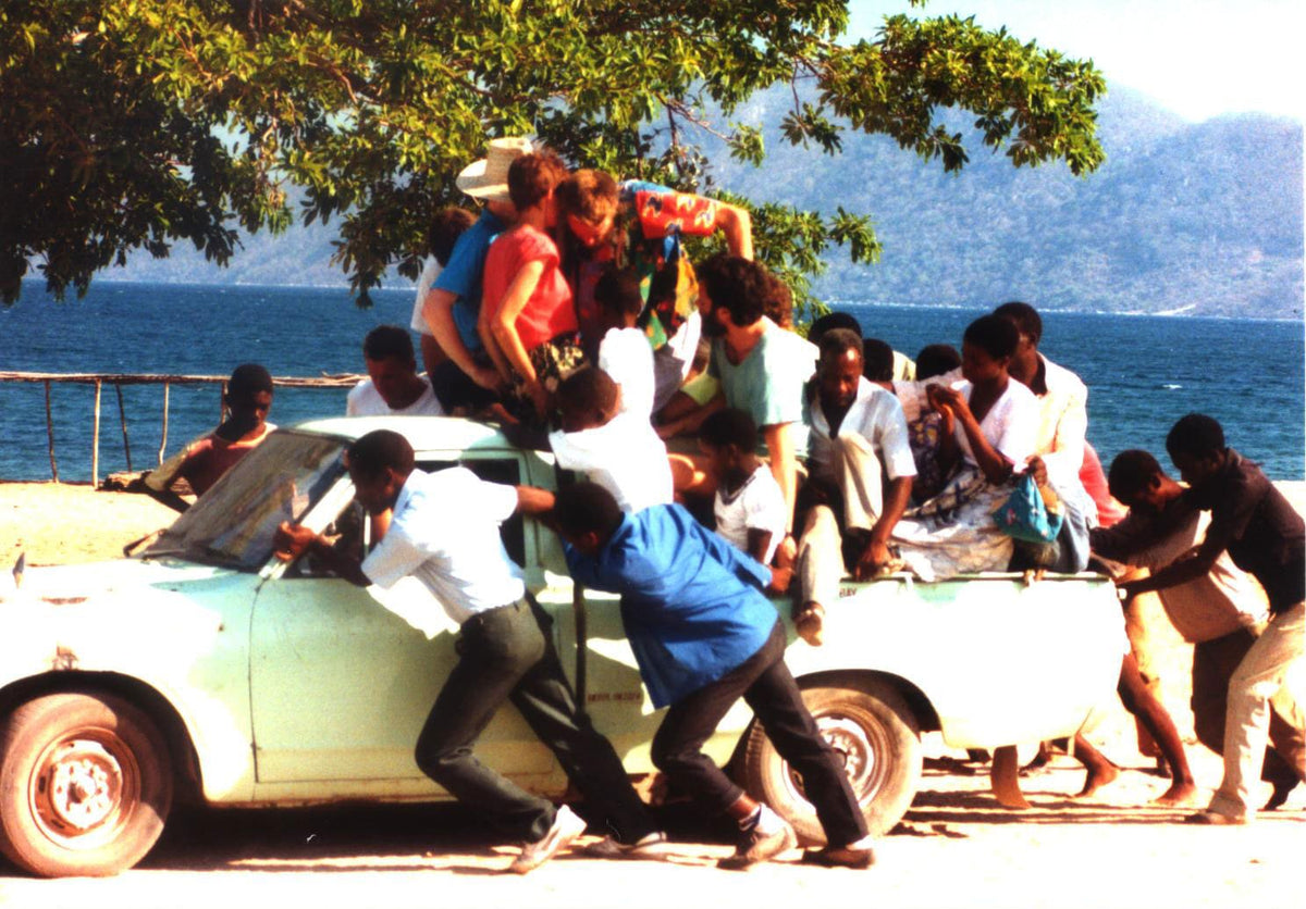 A group of people crowded onto and around a light-colored vintage car on a beach, with ocean water, trees, and mountains in the background.