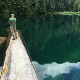 Craig Holt from behind standing on a log in the middle of a lake surrounded by evergreen forests